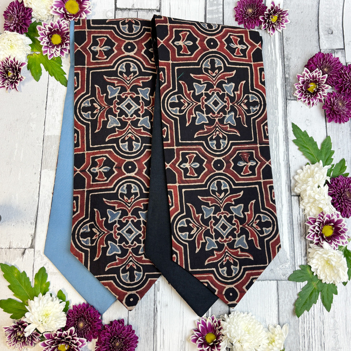 Two cravats with ornate geometric patterns, one in black with red, beige, and gray motifs, and the other in light blue, arranged on a white wood background with decorative flowers.
